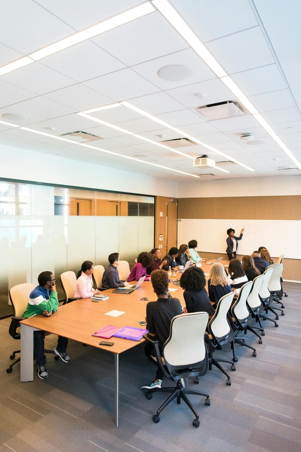 Large group of about twenty people sitting around a conference table watching a presentation. One person is presenting and pointing to a whiteboard.