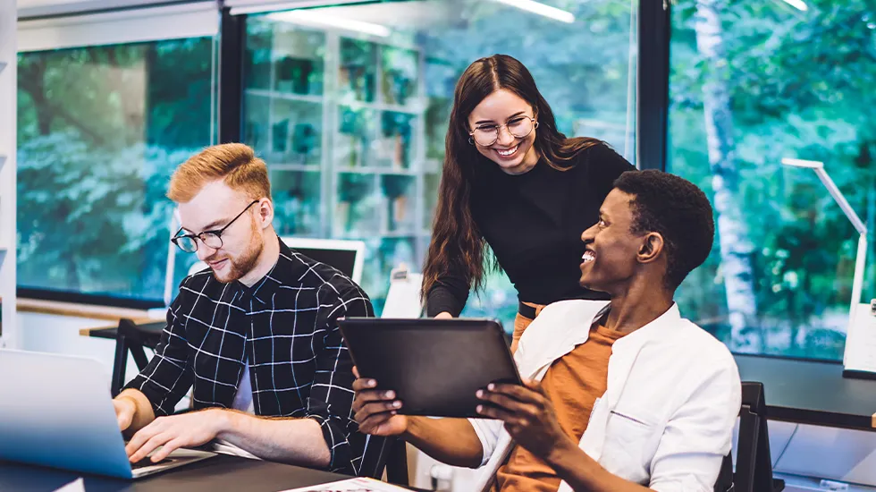 Three colleagues smiling in an office. One is showing another something on an iPad - both are smiling.