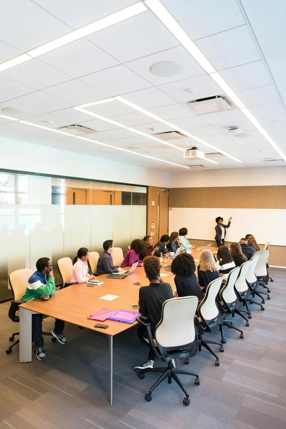 Large group of about twenty people sitting around a conference table watching a presentation. One person is presenting and pointing to a whiteboard.